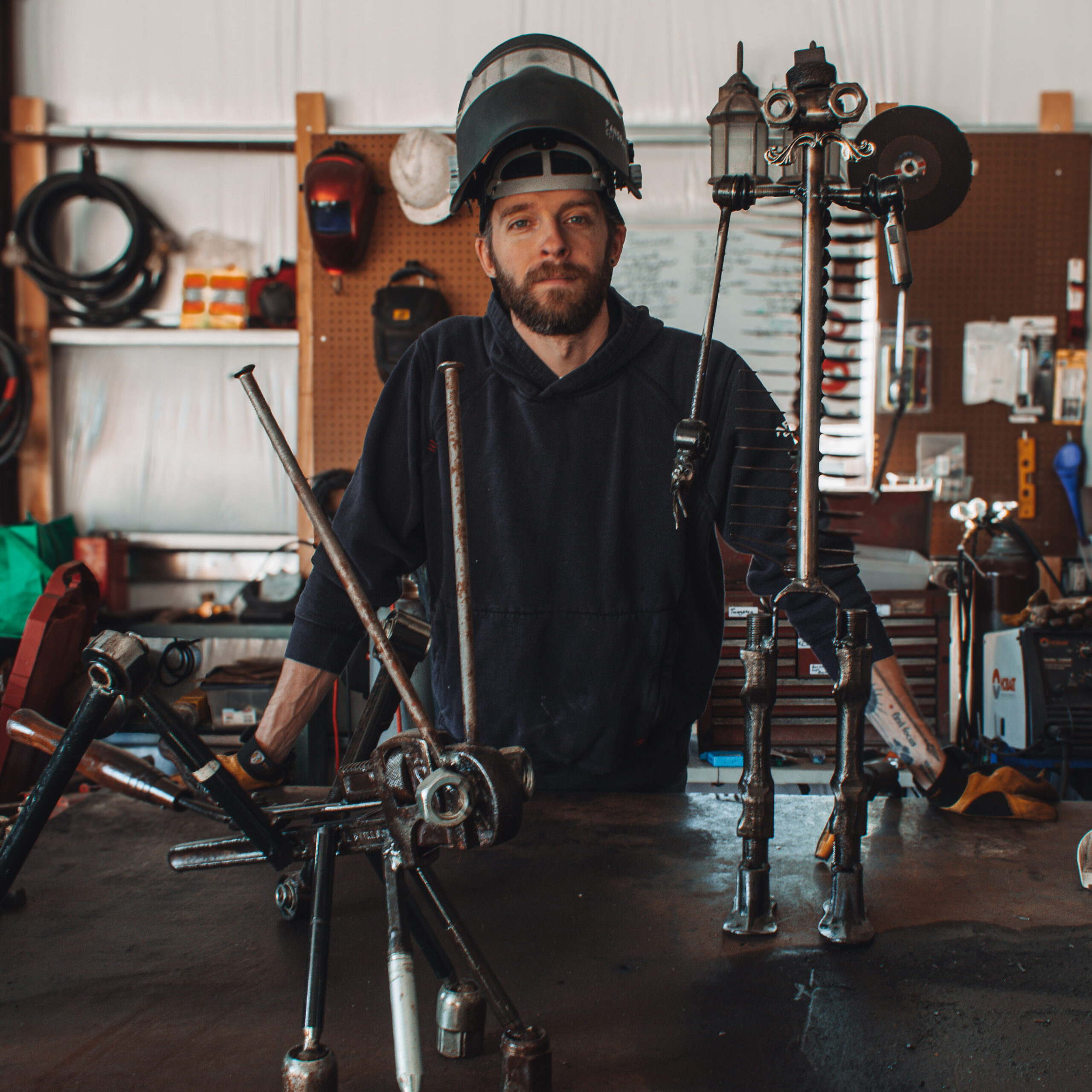 Metal artist Ryan standing at a workbench inside a workshop, wearing a raised welding helmet and hoodie, with two welded metal sculptures in front of him—one resembling a grasshopper and the other a tall humanoid figure—surrounded by tools and industrial equipment.