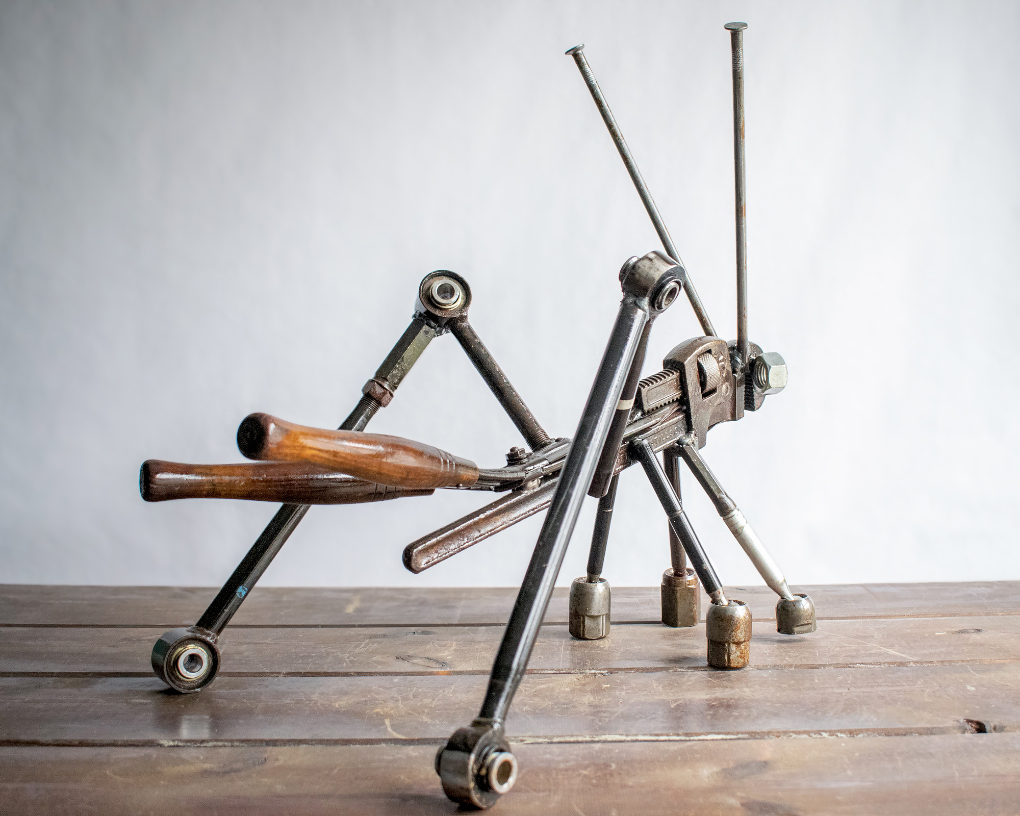 Angled view of reclaimed metal grasshopper sculpture with pipe wrench body and long steel legs.