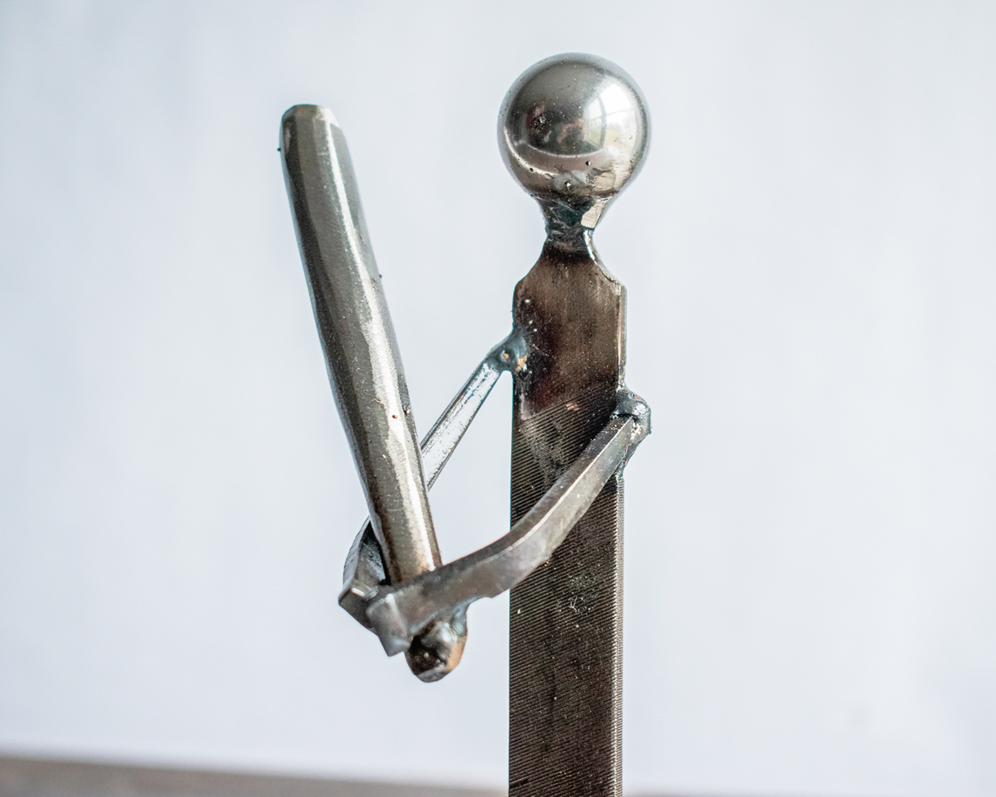 Close-up of welded arms, bat, and metal file body on a baseball player sculpture.