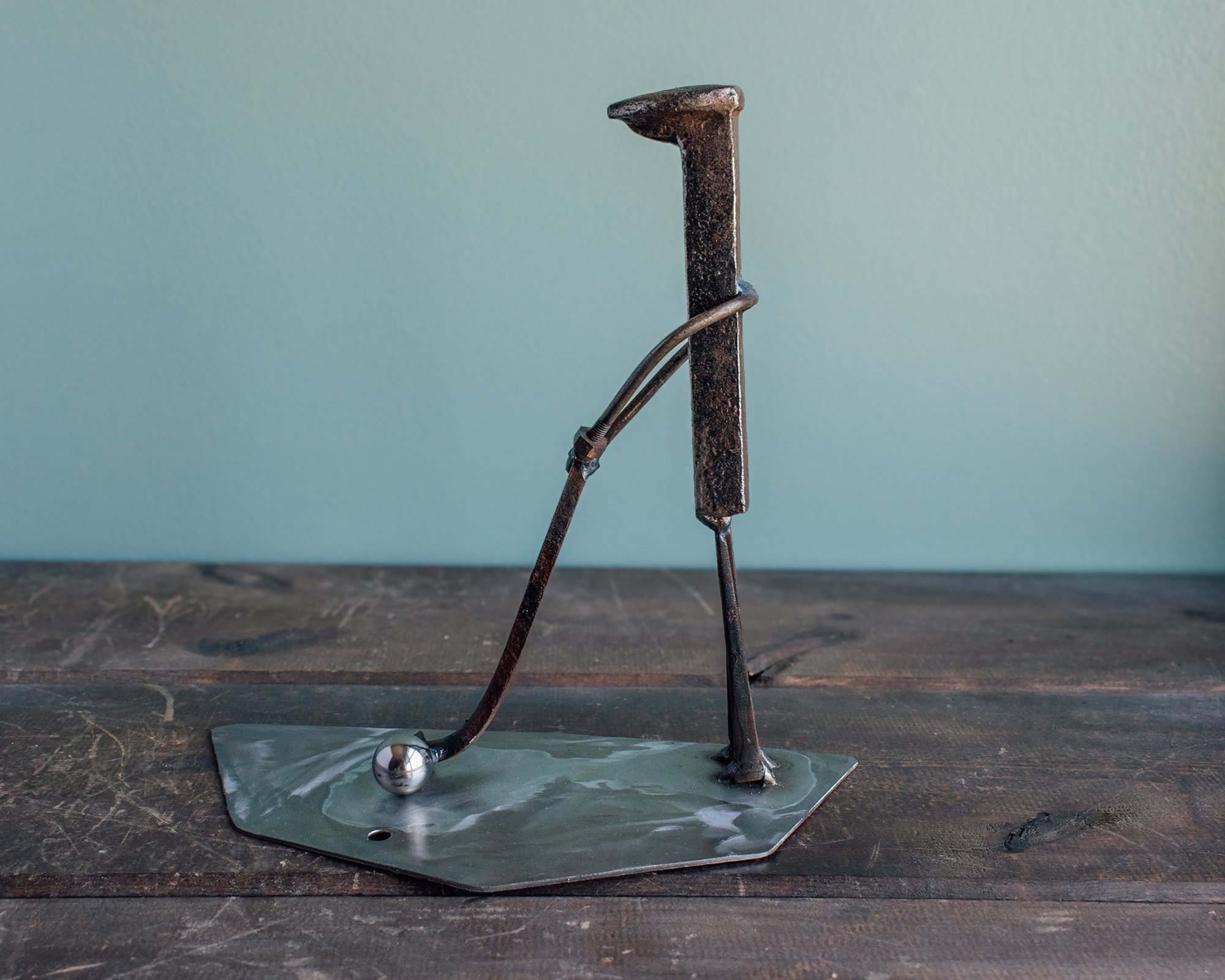 Welded metal golfer sculpture made from a railroad spike, square-head nails, and a ball bearing golf ball.