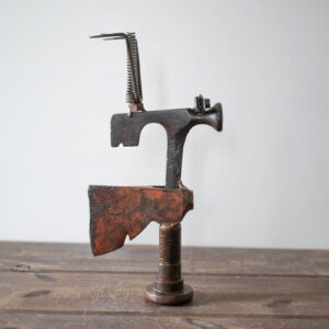 Side profile of a welded metal hatchet sculpture with a raw axe head, large bolt base, and garden rake detail.